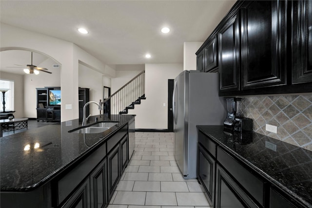 kitchen with sink, decorative backsplash, ceiling fan, dark stone countertops, and an island with sink