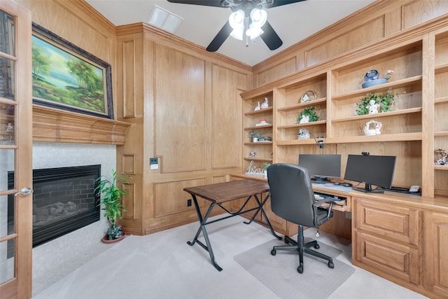 carpeted office with built in desk, ceiling fan, wood walls, and a tiled fireplace