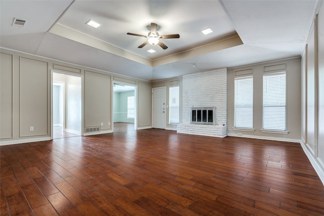 unfurnished living room with ceiling fan, wood-type flooring, a brick fireplace, and a tray ceiling