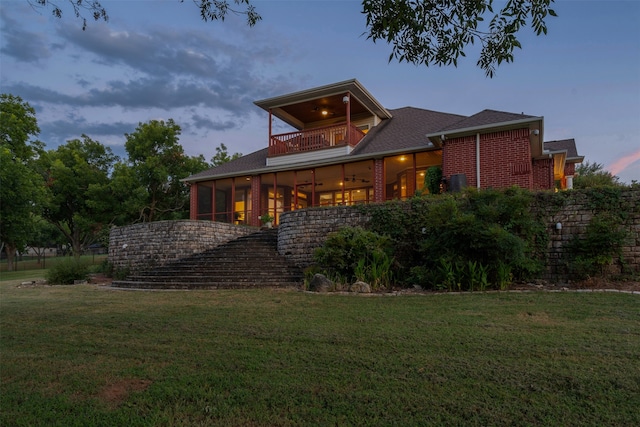 back house at dusk with ceiling fan, a balcony, a lawn, and a sunroom