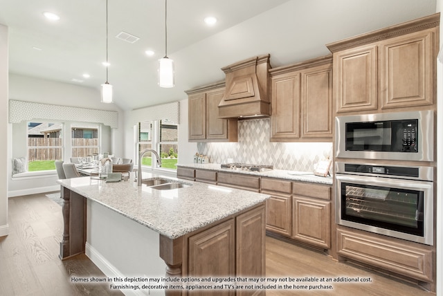 kitchen with stainless steel appliances, a center island with sink, light stone countertops, lofted ceiling, and premium range hood