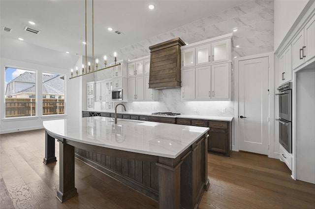 kitchen featuring light stone counters, dark wood-type flooring, hanging light fixtures, a center island with sink, and appliances with stainless steel finishes