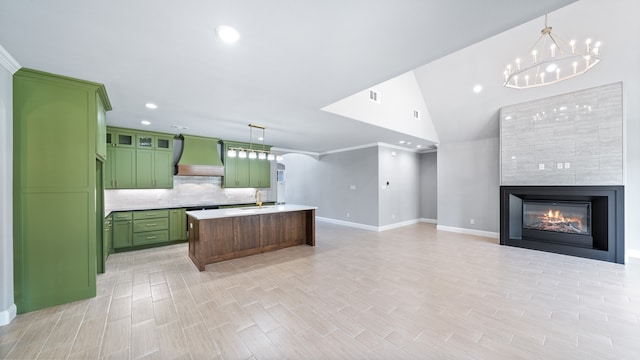 kitchen with a multi sided fireplace, pendant lighting, custom exhaust hood, an island with sink, and green cabinets