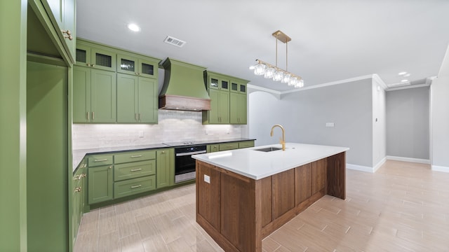 kitchen with stainless steel oven, green cabinets, premium range hood, sink, and a kitchen island with sink