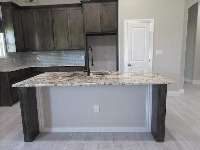 kitchen with light stone countertops, dark brown cabinets, a kitchen island with sink, and sink