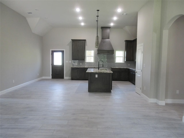 kitchen with custom exhaust hood, light stone countertops, light wood-type flooring, an island with sink, and tasteful backsplash