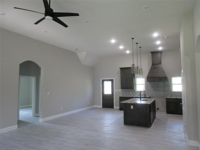 kitchen with hanging light fixtures, premium range hood, light hardwood / wood-style floors, decorative backsplash, and a center island with sink