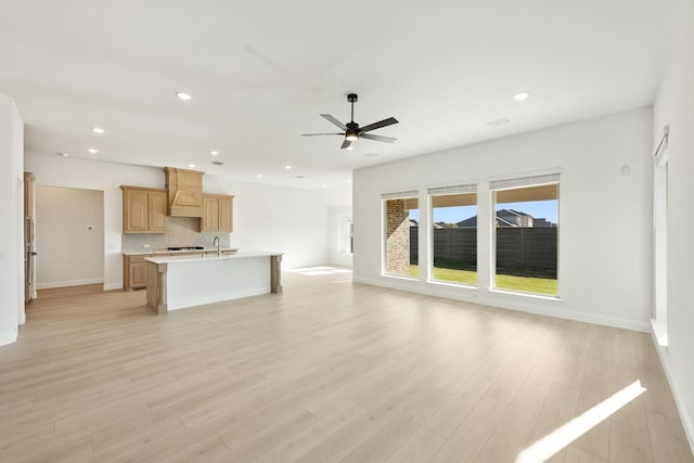 kitchen with backsplash, a kitchen island with sink, light hardwood / wood-style flooring, ceiling fan, and custom range hood