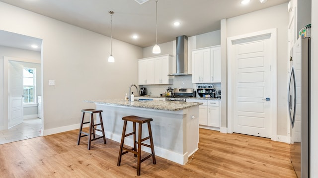 kitchen with stove, stainless steel fridge, a kitchen island with sink, light wood-type flooring, and wall chimney exhaust hood