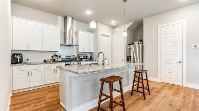 kitchen with stainless steel appliances, wall chimney range hood, a kitchen island with sink, light wood-type flooring, and white cabinetry