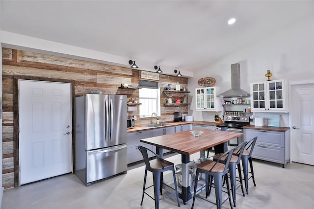 kitchen featuring appliances with stainless steel finishes, wood counters, vaulted ceiling, wall chimney range hood, and sink