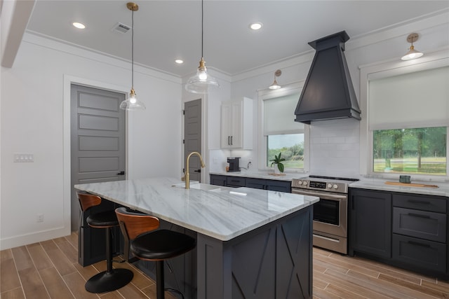 kitchen featuring stainless steel electric stove, a kitchen island with sink, premium range hood, and light wood-type flooring