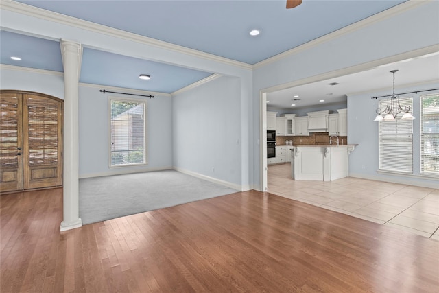 unfurnished living room featuring ornate columns, sink, light hardwood / wood-style floors, crown molding, and an inviting chandelier