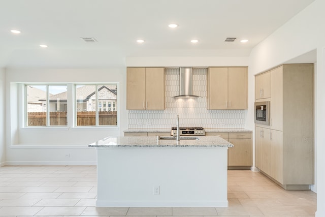 kitchen with wall chimney range hood, backsplash, a center island with sink, light brown cabinetry, and light stone counters