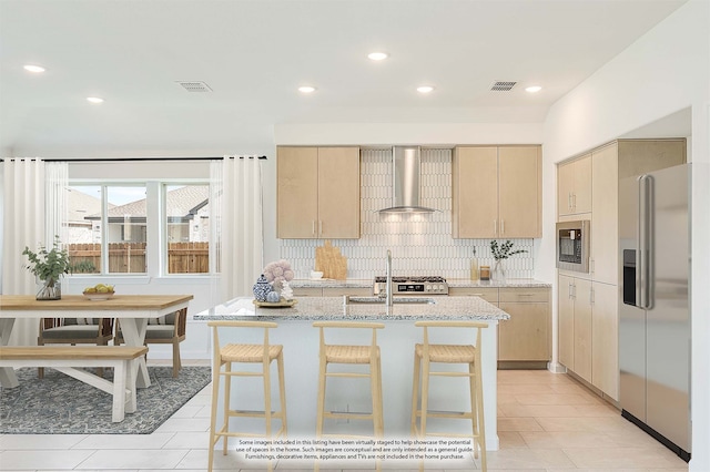 kitchen with stainless steel appliances, wall chimney range hood, an island with sink, and light brown cabinets