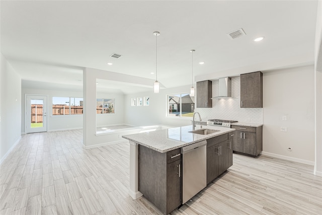 kitchen featuring wall chimney exhaust hood, stainless steel dishwasher, decorative light fixtures, a center island with sink, and light wood-type flooring