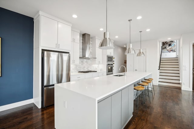 kitchen with tasteful backsplash, wall chimney range hood, dark hardwood / wood-style flooring, appliances with stainless steel finishes, and a center island with sink