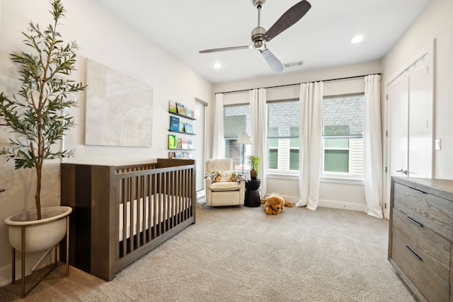 bedroom with a crib, light colored carpet, multiple windows, and ceiling fan