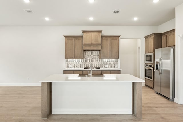 kitchen featuring stainless steel appliances, an island with sink, light hardwood / wood-style floors, and decorative backsplash