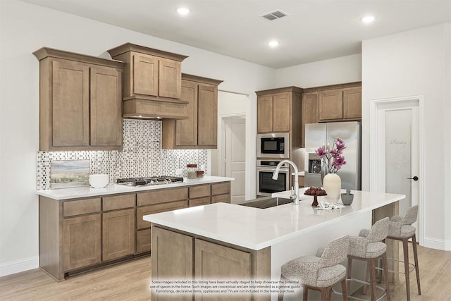 kitchen featuring sink, light wood-type flooring, stainless steel appliances, a kitchen island with sink, and decorative backsplash