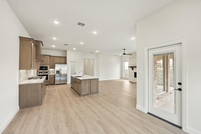 kitchen with an island with sink, a large fireplace, backsplash, stainless steel appliances, and light wood-type flooring