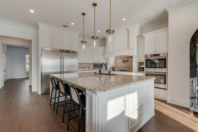 kitchen with light stone counters, white cabinets, a large island with sink, decorative light fixtures, and built in appliances