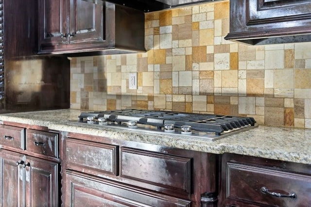 kitchen with stainless steel gas stovetop, backsplash, exhaust hood, light stone counters, and dark brown cabinetry