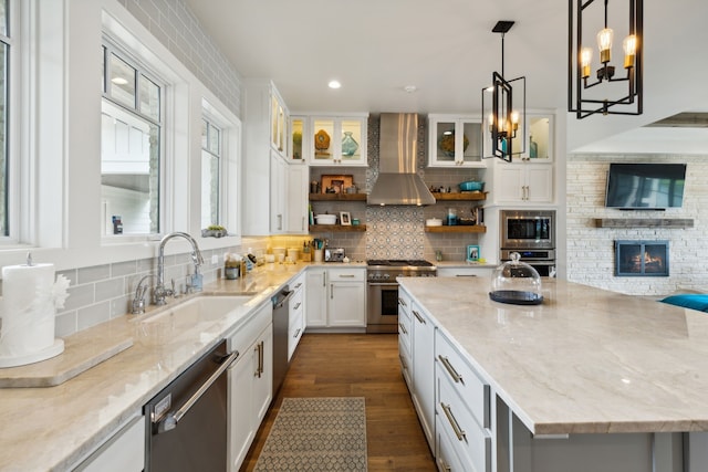 kitchen with wall chimney range hood, pendant lighting, stainless steel appliances, decorative backsplash, and a stone fireplace