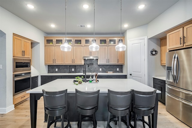 kitchen with stainless steel appliances, light brown cabinetry, an island with sink, and backsplash