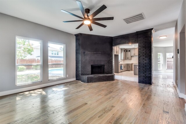 unfurnished living room with decorative columns, visible vents, light wood-style floors, a ceiling fan, and a brick fireplace