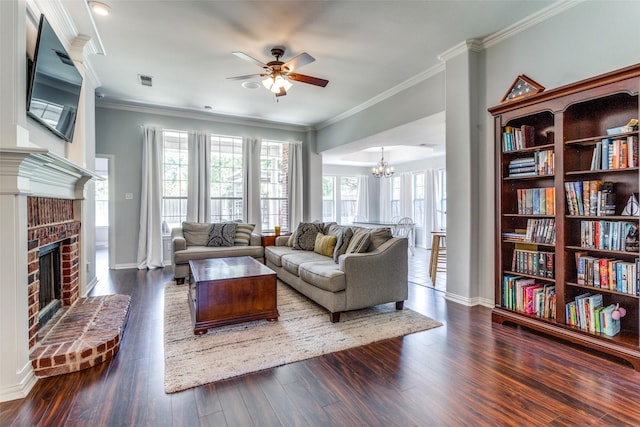 living room with a brick fireplace, dark wood-type flooring, ornamental molding, and ceiling fan with notable chandelier