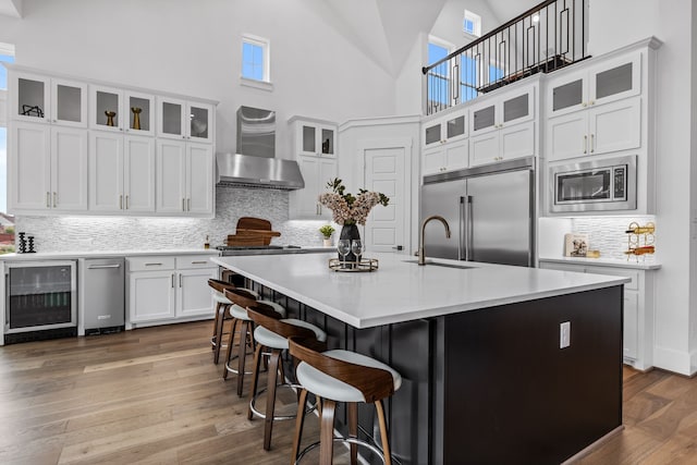 kitchen with wall chimney exhaust hood, built in appliances, an island with sink, beverage cooler, and white cabinets