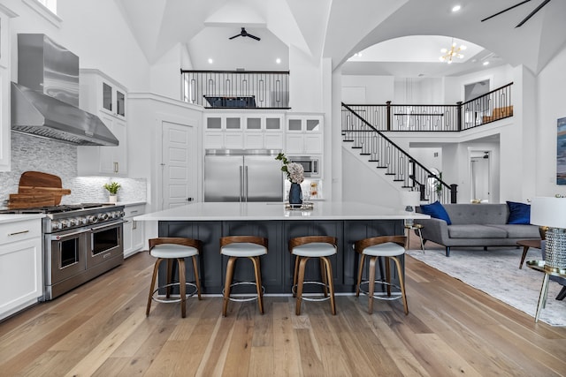 kitchen with wall chimney range hood, a kitchen island with sink, built in appliances, and white cabinets