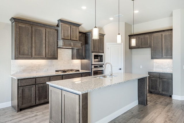 kitchen with light stone countertops, light wood-type flooring, stainless steel appliances, a kitchen island with sink, and hanging light fixtures