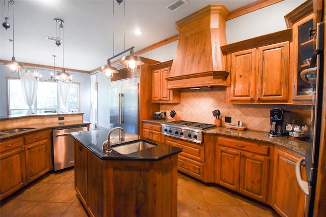 kitchen with hanging light fixtures, appliances with stainless steel finishes, sink, premium range hood, and an island with sink