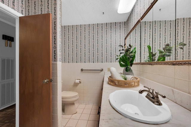 bathroom with wallpapered walls, wainscoting, toilet, vanity, and tile walls