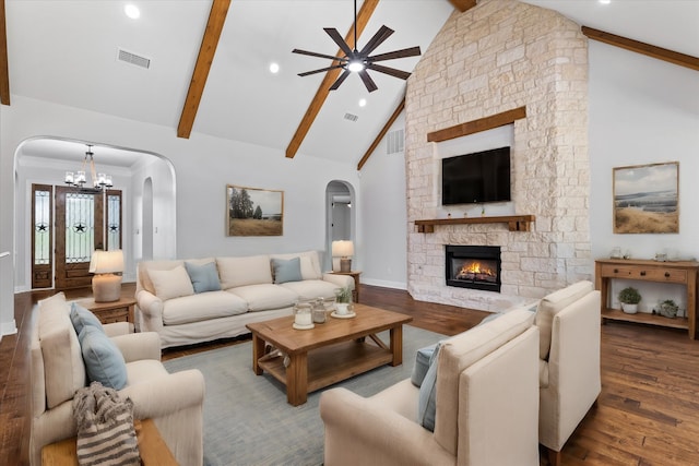 living room with ceiling fan with notable chandelier, high vaulted ceiling, beam ceiling, a stone fireplace, and dark wood-type flooring