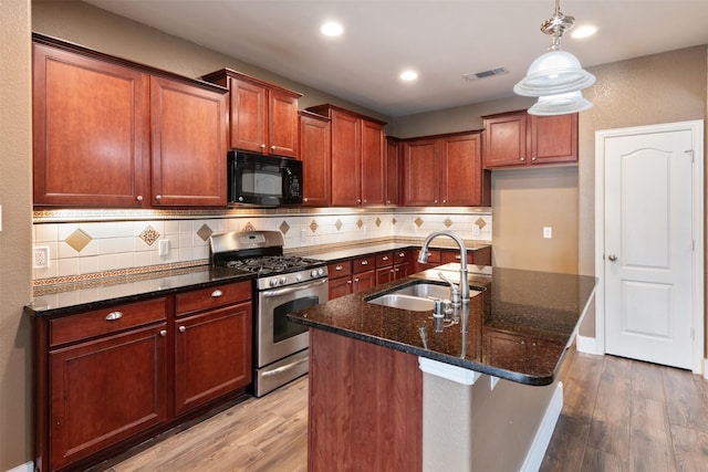 kitchen with sink, stainless steel range with gas cooktop, dark stone countertops, a center island with sink, and hardwood / wood-style flooring