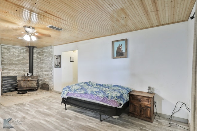 bedroom featuring ceiling fan, a wood stove, wooden ceiling, brick wall, and light hardwood / wood-style floors