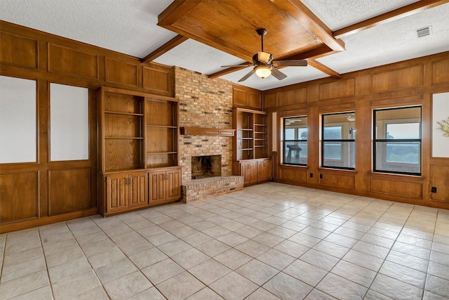 unfurnished living room with built in features, beam ceiling, wooden walls, a textured ceiling, and a brick fireplace