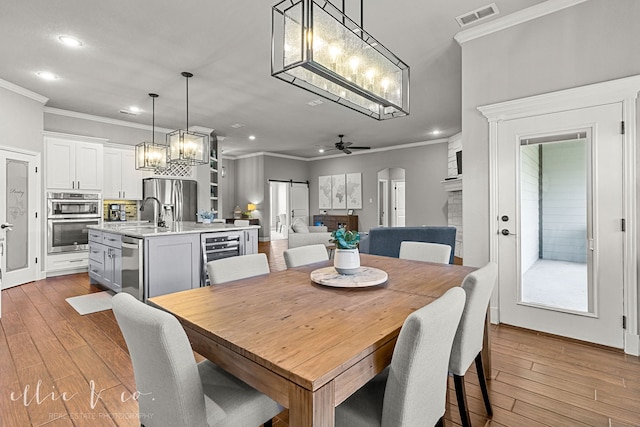 dining space with wine cooler, ceiling fan with notable chandelier, light hardwood / wood-style floors, sink, and ornamental molding