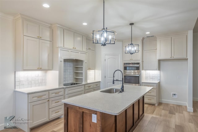 kitchen with an island with sink, stainless steel double oven, white cabinetry, pendant lighting, and a sink