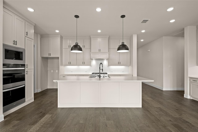 kitchen featuring black microwave, hanging light fixtures, a center island with sink, dark hardwood / wood-style flooring, and white cabinets