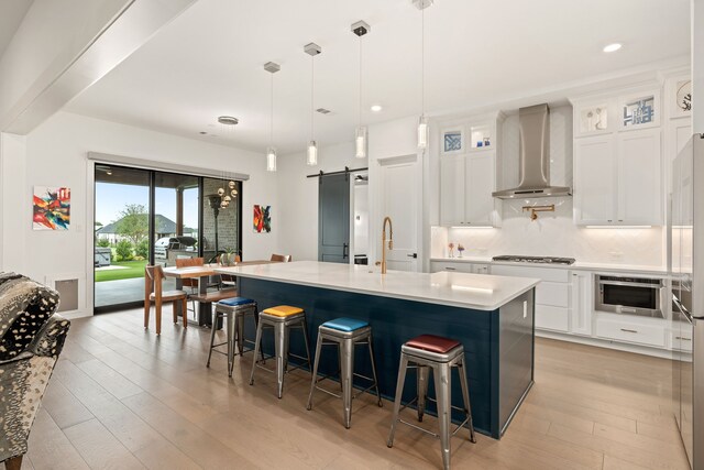 kitchen with a barn door, a center island with sink, hanging light fixtures, white cabinets, and wall chimney exhaust hood