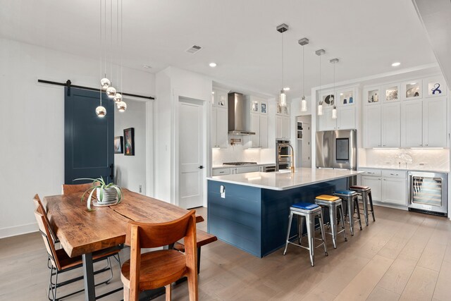 kitchen featuring white cabinets, wall chimney exhaust hood, decorative light fixtures, an island with sink, and a barn door