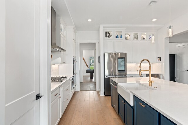 kitchen featuring stainless steel appliances, blue cabinetry, wall chimney range hood, pendant lighting, and white cabinets