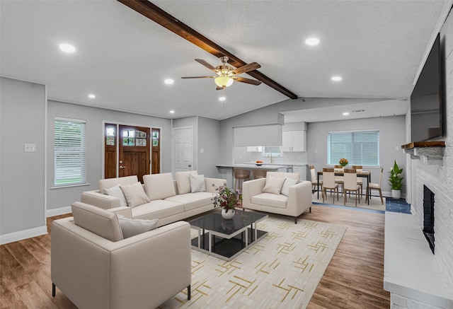 living room with ceiling fan, a fireplace, vaulted ceiling with beams, light hardwood / wood-style flooring, and a textured ceiling