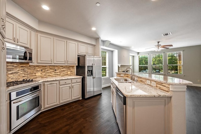 kitchen with sink, a kitchen island with sink, backsplash, stainless steel appliances, and light stone countertops