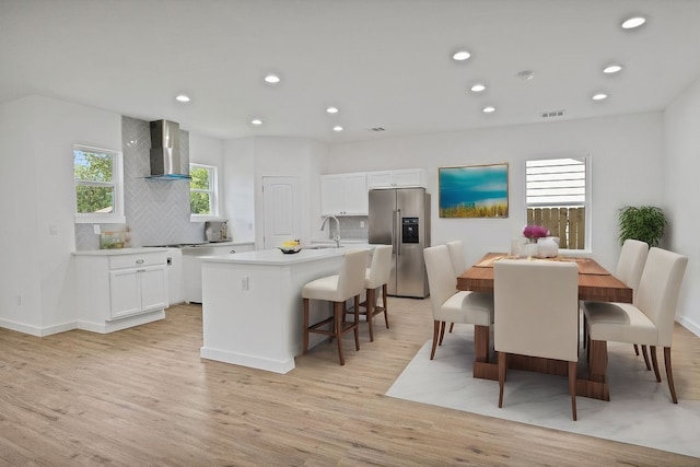 kitchen featuring stainless steel refrigerator with ice dispenser, wall chimney exhaust hood, sink, a center island with sink, and white cabinets