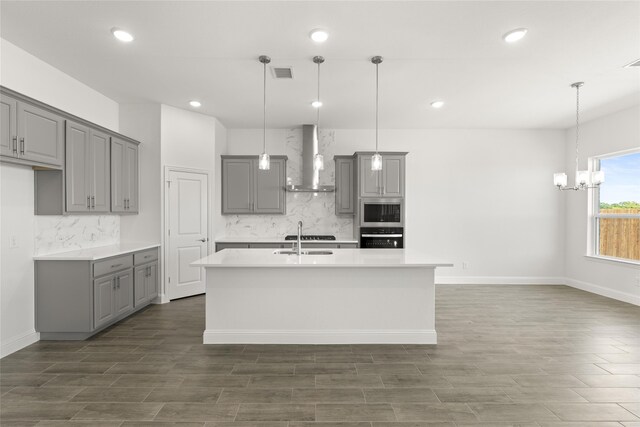 kitchen with a center island with sink, stainless steel appliances, decorative light fixtures, gray cabinets, and wall chimney exhaust hood